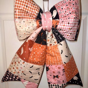 May include: A large, quilted fabric bow featuring a patchwork design with various Halloween-themed patterns including ghosts, bats, pumpkins, black cats, spider webs, and potion bottles in shades of orange, pink, black, and cream, tied with a pink centre.