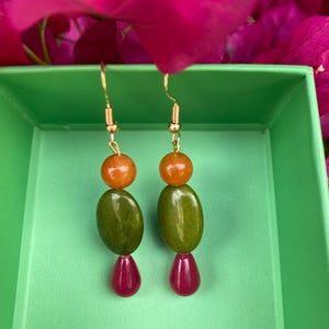 Può includere: Orecchini fatti a mano con ganci color oro. Ogni orecchino presenta una sfera arancione, un ovale verde e una perla a goccia rosso scuro. Gli orecchini sono esposti in una scatola verde, con fiori rosa sullo sfondo.