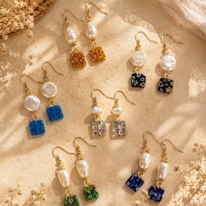 May include: A collection of handmade earrings with gold-colored hooks. The earrings feature white pearls and colorful square beads with floral patterns. The beads are in various colors, including blue, green, orange, and black. The earrings are displayed on a beige surface.