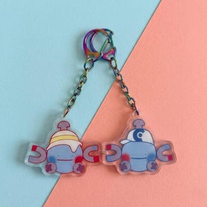 Può includere: Un portachiavi in acrilico trasparente con due personaggi dei cartoni animati blu con magneti, collegati da una catena e una chiusura color arcobaleno. Un personaggio indossa un cappello con una striscia gialla e bianca, l'altro un berretto blu e bianco.