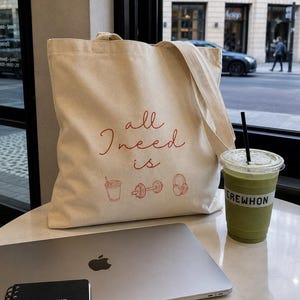 May include: A beige canvas tote bag featuring the red text "all I need is" above red line art icons of a drink, a dumbbell, and headphones. Next to it sits a green smoothie in a clear plastic cup labeled "EREWHON" and a silver laptop.