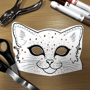 May include: A white paper cat mask with black outlines and orange spots, alongside scissors, a hole punch, and markers. The mask features eye holes and a smiling mouth, perfect for a craft project.