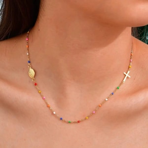 May include: Gold necklace with a series of colourful beads and a cross pendant. The necklace also includes a medallion. The beads are in various colours, including red, orange, yellow, green, blue, and pink.