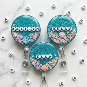 May include: Three round, teal glitter badge reels with the names "KAYTLYN", "BELLA", and "ELLA" spelled out in white letters. Each reel features miniature seashells, starfish, and coral. The reels have a clear plastic strap and a silver metal clip.