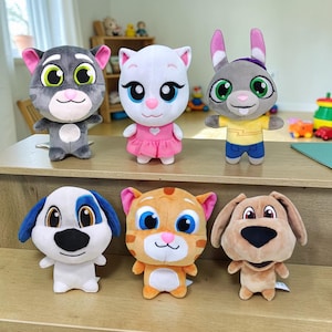 Puede incluir: Una colección de seis peluches que representan personajes de dibujos animados. La fila superior incluye un gato gris, una gata blanca con vestido rosa y un conejo gris con camisa amarilla. La fila inferior muestra un perro blanco y azul, un gato atigrado naranja y un perro color canela.