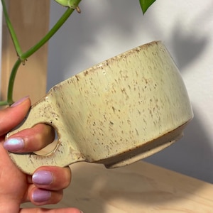 May include: A ceramic mug with a pale yellow, speckled glaze and a wide, flat handle featuring a circular cutout. The mug has a slightly tapered body with a visible base rim, showcasing a rustic, textured finish throughout the exterior surface.
