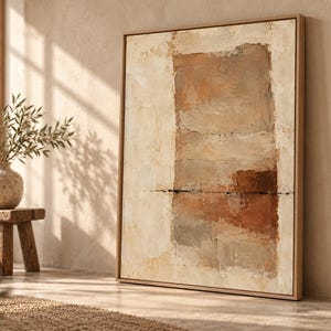 May include: A rectangular canvas painting featuring an abstract composition of layered earth tones including cream, beige, tan, and deep brown. The artwork is enclosed in a light-colored wooden frame and displays a textured, block-like paint application.