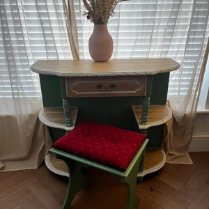Op de afbeelding: Een groene en houten kaptafel met een lade, een bijpassende kruk met een rood kussen en een vaas met gedroogde bloemen. De tafel heeft een licht houten blad en een groene basis. De kruk heeft een rood kussen met witte stippen.