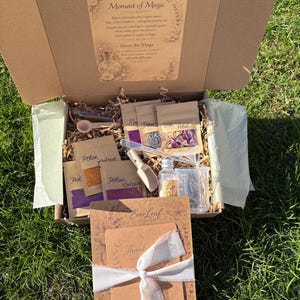 Puede incluir: Una caja de cartón abierta llena de varios artículos, incluyendo pequeños paquetes etiquetados como "Potion Colour" y "Sea Dreams with Petals". También se ven una pequeña botella de vidrio y una cuchara de madera. La caja está atada con una cinta blanca e incluye el texto "A Little Moment of Magic".