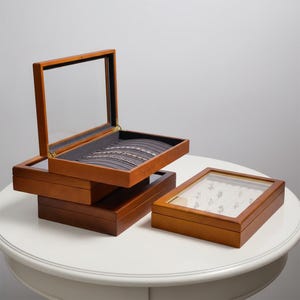 May include: Wooden jewelry boxes with glass lids. The boxes are various sizes and shapes, with a warm brown finish. One box is open, revealing a gray interior with jewelry. Another box contains rings.