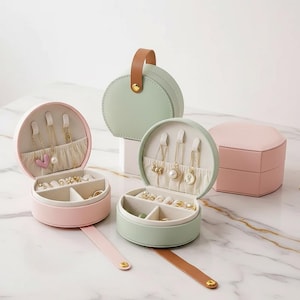 May include: A selection of pastel-coloured jewellery boxes. The open boxes display compartments for rings and necklaces. The boxes are round and have a handle or strap. The colours are pink, green, and light brown.