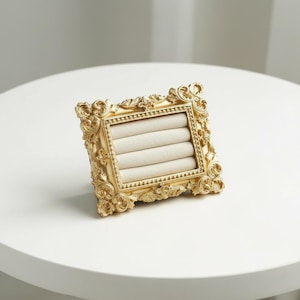 May include: A gold-colored, ornate jewelry display stand with a square shape. The stand features a detailed, scrollwork design around the frame and a beige, padded interior with multiple slots for rings. The stand is on a white surface.