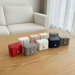 Puede incluir: Una colección de pequeñas joyeros cuadrados en varios colores, incluyendo rojo, gris, blanco, rosa y azul marino. Cada caja tiene una superficie texturizada, un cierre de botón dorado y un diseño compacto, perfecto para guardar objetos pequeños. (Medidas en cm)