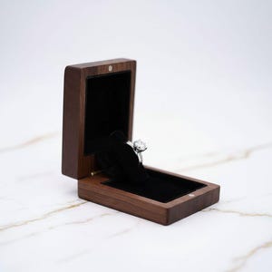 Può includere: Un anello di fidanzamento con diamanti esposto in un portagioie in legno scuro aperto, con interno in velluto nero. L'anello è posto su un supporto in velluto nero, che ne evidenzia la brillantezza. Il portagioie è su una superficie di marmo bianco.