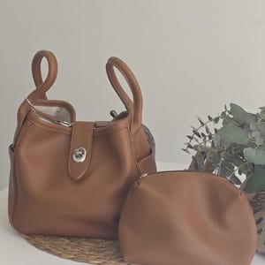 Può includere: Una borsa color cuoio con due manici arrotondati, una chiusura a girello in metallo argentato su una linguetta centrale e una pochette con cerniera abbinata. La borsa presenta una superficie testurizzata e un design aperto con fodera chiara.