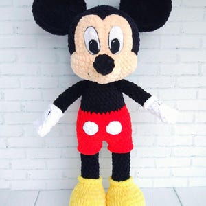 May include: A handmade crochet Mickey Mouse doll. The doll features a black head with large round ears, a tan face, and a black nose. It wears red shorts with white polka dots, black legs, yellow shoes, and white gloves.