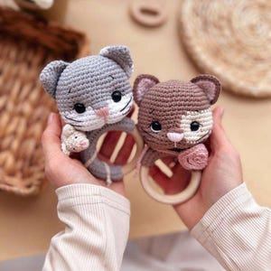 May include: Two crocheted cat toys, one gray and one brown, each holding a wooden ring. The gray cat has a small crocheted mouse attached. Both toys have black eyes, pink noses, and white accents. The toys are handmade.