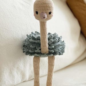 May include: A handmade crochet ostrich doll with a beige head, neck, and legs, and a blue-grey ruffled body. The ostrich has small black eyes, pink cheeks, and a pink beak. The doll is standing on a white surface.