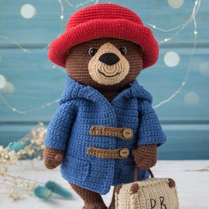 Puede incluir: Oso de ganchillo marrón con sombrero rojo y abrigo azul con cierres de madera. El oso sostiene una pequeña maleta beige con las iniciales "P.B." en ella. Artículo hecho a mano.