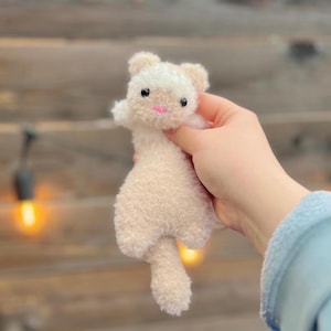 May include: A small, plush cat toy in cream and white, held in a hand. The toy has a fluffy texture, with a white face and ears, black eyes, and a pink mouth. The background is a blurred wooden surface.