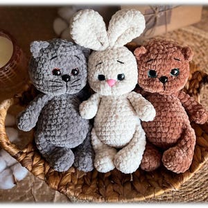 Puede incluir: Tres animales de peluche de ganchillo hechos a mano: un gato gris, un conejito blanco con nariz rosa y un oso pardo. Están sentados en una cesta de mimbre. Los juguetes tienen ojos negros y están hechos de hilo suave y afelpado.