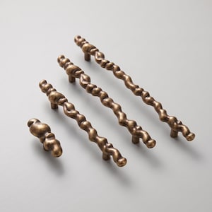 Peut inclure: Un ensemble de quatre poignées de placard avec une finition bronze métallique, présentant une texture irrégulière, organique et ondulée. La collection comprend un petit bouton et trois poignées allongées de longueurs variées, chacune avec une surface bosselée.