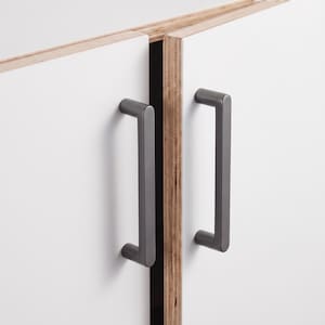May include: Two matte dark gray metal cabinet handles mounted on white cabinet doors. The doors feature exposed plywood edges with visible wood grain layers, creating a contrast between the smooth white surfaces and the textured wooden sides.