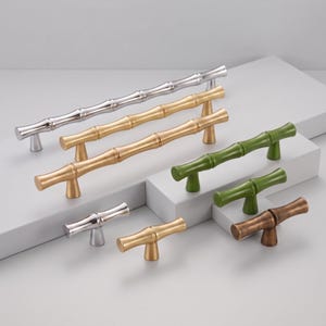 May include: A collection of cabinet pulls and knobs designed with a bamboo-inspired texture. The set includes various lengths and finishes, featuring polished silver, brushed gold, matte green, and antique bronze tones with two mounting posts.