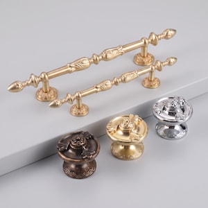 Peut inclure: Deux poignées de placard en métal doré allongé avec des embouts décoratifs et des détails centraux, accompagnées de trois boutons de placard ronds en bronze foncé, or brossé et chrome poli, chacun avec un motif floral sur le dessus.