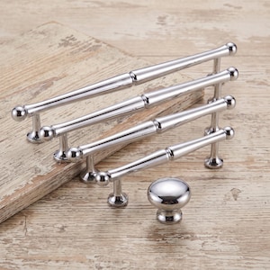 May include: A collection of four polished chrome cabinet pulls of varying lengths and one matching round cabinet knob. Each pull features a slender, cylindrical bar design with rounded ball-shaped ends and two sturdy mounting posts for attachment.