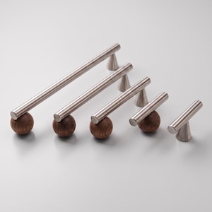 Op de afbeelding: Een set van vijf kastgrepen in verschillende lengtes, elk bestaande uit een horizontale staaf van geborsteld metaal op een ronde, donkerbruine houten basis en een klein conisch metalen accentstuk, diagonaal gerangschikt op grootte.