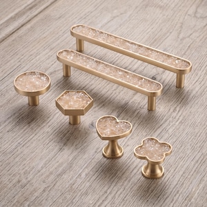 Op de afbeelding: Een set van zes goudkleurige metalen kastbeslagstukken, waaronder twee lange rechthoekige handgrepen, een ronde knop, een zeshoekige knop, een hartvormige knop en een klavervormige knop, allemaal ingelegd met heldere, gebroken kristalaccenten.