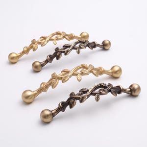 May include: Four metal cabinet pulls arranged in a staggered formation. Each handle features a curved, vine-like structure adorned with small leaves and finished with a spherical knob at both ends. Two pulls are gold-toned and two are dark bronze.