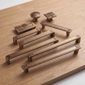 May include: A collection of bronze-toned metal cabinet hardware including various lengths of rectangular drawer pulls, a square knob, a round knob, and a rectangular recessed pull, all featuring a consistent geometric Greek key pattern.