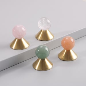 Puede incluir: Cuatro objetos decorativos, cada uno compuesto por una piedra preciosa esférica y lisa montada sobre una base cónica de metal dorado cepillado. Las esferas presentan tonos rosa pálido, blanco translúcido, verde suave y color melocotón.