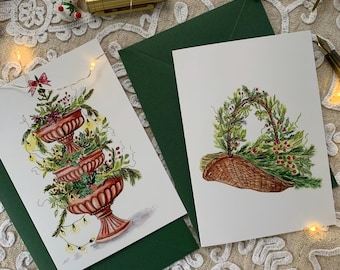 May include: Two white greeting cards featuring watercolour illustrations. One card shows a three-tiered terracotta urn filled with evergreen branches, berries, and string lights. The other card displays a woven basket overflowing with greenery and red berries.