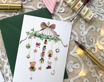 May include: A white greeting card featuring a watercolour illustration of a Christmas-themed mobile with a plaid bow, pine branches, and hanging ornaments including a teddy bear, a stocking, stars, a bell, and blocks spelling out the word BABY.