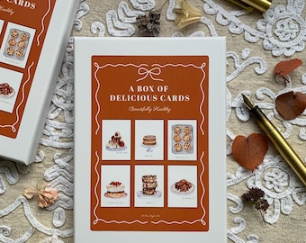 May include: A white rectangular box with a burnt orange centre panel featuring the text A BOX OF DELICIOUS CARDS and Beautifully Healthy. The box displays six illustrations of desserts including cakes, cookies, and pastries in a grid layout.