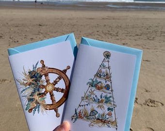 May include: Two white greeting cards with light blue envelopes. One card features a brown wooden ship wheel adorned with starfish and blue foliage. The other card displays a triangular driftwood structure decorated with seashells, beach items, and blue greenery.