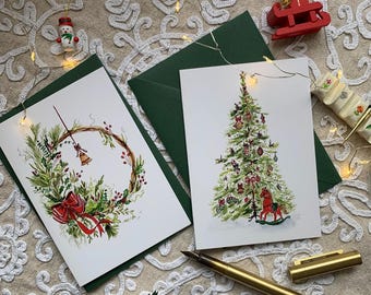 May include: Two white greeting cards with watercolour illustrations. One card features a green wreath with a red bow and a gold bell. The other card displays a decorated Christmas tree with a red rocking horse at the base. Both cards have dark green envelopes.
