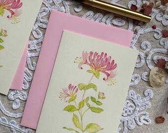 May include: A cream-coloured greeting card featuring a watercolour illustration of a honeysuckle plant with pink and yellow flowers and green leaves. The word honeysuckle is written in cursive at the bottom. A gold fountain pen rests nearby.