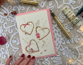 May include: A cream-coloured greeting card featuring a watercolour illustration of three heart-shaped wreaths made of twigs, each adorned with small pink flowers, ribbons, and delicate botanical accents, set against a pink cardstock border.