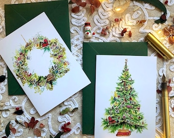 May include: Two white greeting cards with watercolour illustrations. One card features a circular wreath made of greenery and small objects, while the other displays a decorated Christmas tree standing on a stack of books. Both cards are paired with dark green envelopes.