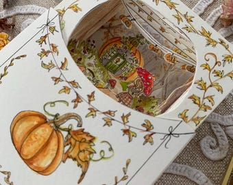 May include: A white paper craft box featuring a hand-drawn orange pumpkin and autumn leaves on the exterior. The top has a circular cutout revealing a multi-layered 3D diorama inside, depicting a small house, mushrooms, and hanging pumpkin lanterns.