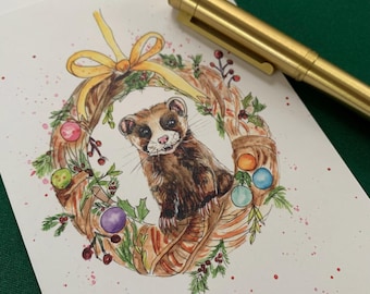 May include: A watercolour illustration on white paper featuring a brown ferret centred within a festive wreath decorated with a yellow ribbon, green pine needles, red berries, and colourful ornaments, accompanied by a metallic gold pen.