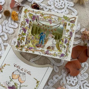 May include: A layered paper shadow box card featuring a watercolour illustration of a couple in a vineyard with a banner reading JUST MARRIED. A separate card shows two wine glasses and the text Mr & Mrs in a decorative script font.