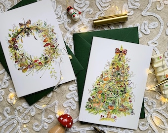 May include: Two white greeting cards featuring watercolour illustrations. One card displays a circular wreath with forest animals and foliage, while the other shows a Christmas tree decorated with woodland creatures, mushrooms, and a red patterned bow.