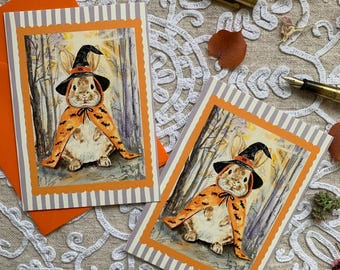 May include: Two rectangular greeting cards featuring a watercolour illustration of a rabbit wearing a black witch hat and an orange cape with black bat patterns. The cards have a scalloped orange border and a grey and white striped background.