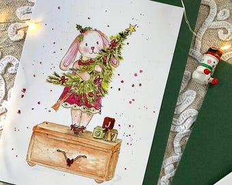 May include: A greeting card featuring a watercolour illustration of a light brown rabbit wearing a pink dress, standing on a wooden chest while holding a decorated Christmas tree. Two small blocks with the letters J and O are visible on the chest.
