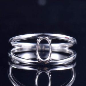 Puede incluir: Un anillo de metal plateado con un diseño de banda dividida que converge en un engaste central ovalado con garras. El engaste está vacío, diseñado para sostener una piedra preciosa, y la banda tiene un acabado superficial pulido y brillante.